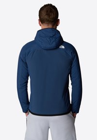 Mörkblå huva-jacka med en texturerad yta, med en logotyp på den övre vänstra ryggen, och ett svart ärmskydd, bärs över ljusa shorts.