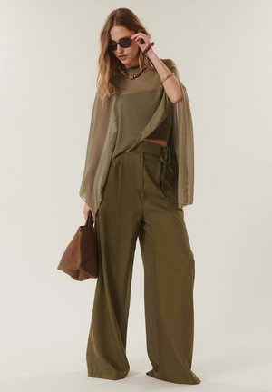 Femme en blouse transparente vert olive et pantalon à jambes larges, portant des lunettes de soleil, tenant un sac à main tressé marron et accessoirisée de bracelets et d'un collier.