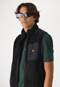 Black fleece vest with zip front, high collar, and a solid black pocket. Worn over a green shirt, paired with black sunglasses.