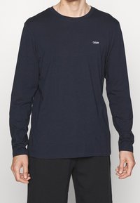 Navyblaues T-Shirt mit langen Ärmeln aus weichem Stoff, ausgestattet mit einem runden Ausschnitt und einem kleinen weißen Logo auf der linken Brustseite.