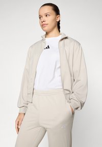 Beige zip-up jakke med en glatt tekstur, båret over en hvit t-skjorte med en svart logo, kombinert med matchende beige bukser.