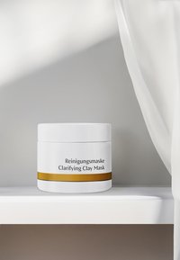 White round container with a gold stripe, labeled "Reinigungsmaske Clarifying Clay Mask." Smooth texture, minimalistic design, placed on a shelf.