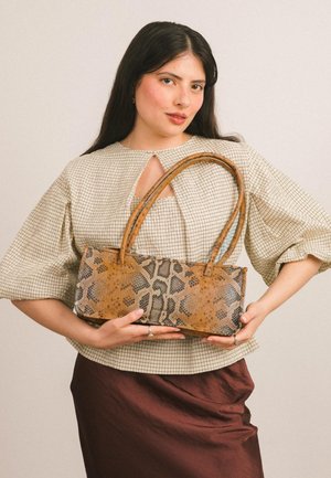 Kobieta o długich ciemnych włosach, ubrana w beżową koszulę w kratę i brązową spódnicę, trzymająca prostokątną torebkę w wzór skóry węża z podwójnymi uchwytami.