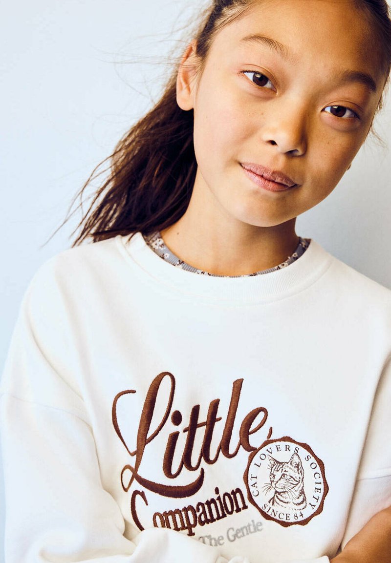 Jeune fille aux longs cheveux foncés portant un sweat-shirt blanc avec le texte "Little Companion Cat Lovers Society" et une illustration de chat.