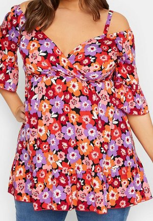 Femme portant un chemisier coloré à fleurs à épaules dénudées avec des fleurs violettes, rouges, oranges et roses, manches et ourlet à volants.