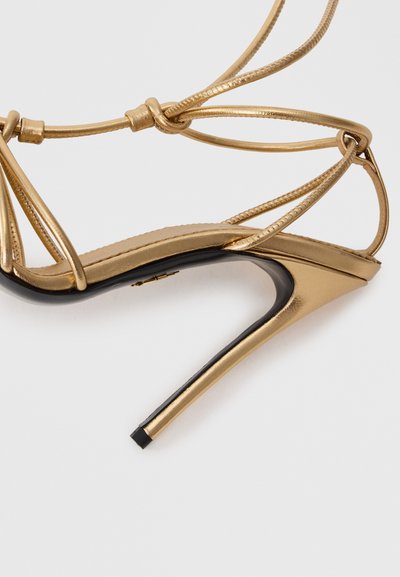 Gouden strappy hoge hak sandaal met een glanzende zwarte zool, met een slank ontwerp, dunne banden en een spitse neus.