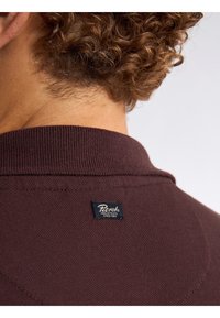 Brązowa dzianinowa koszulka polo z prążkowanym kołnierzykiem i małą czarną metką z białym napisem z tyłu. Gładka faktura z detalicznym ściegiem.