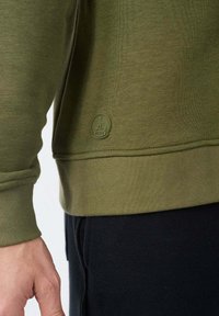 Primer plano de una persona que lleva una sudadera verde oliva con puños acanalados y un pequeño parche redondo de logo cerca del dobladillo, combinada con pantalones negros.