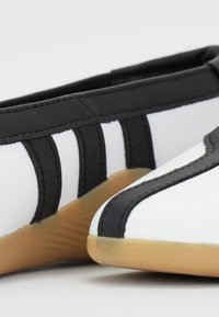 Sapatos brancos e pretos com um cabedal sintético liso e sola de borracha, com bordas curvas e riscas pretas contrastantes de cada lado.