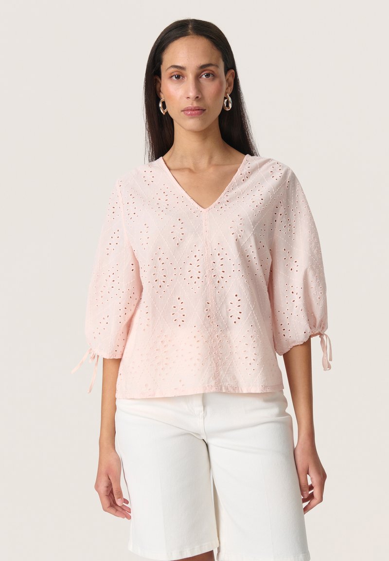 Blusa rosa chiaro con scollatura a V, caratterizzata da motivi floreali intagliati e maniche a sbuffo corte con lacci ai polsi. Indossata con pantaloncini bianchi.