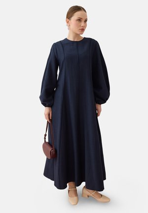 Vrouw draagt een lange marineblauwe jurk met ballonmouwen, beige lage hakken en houdt een kleine bruine handtas vast tegen een effen achtergrond.
