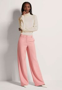 Crème turtlenecktrui gedragen met hooggewelfde, wijde roze broeken. De broek heeft een sluiting met knoop aan de voorkant en een zachte textuur.