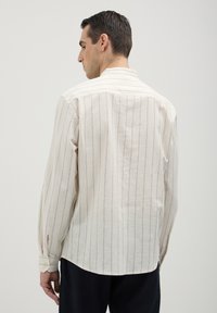 Uomo di spalle che indossa una camicia beige a maniche lunghe con sottili righe verticali e pantaloni scuri, di fronte a uno sfondo chiaro e uniforme.