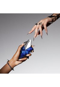 Blaues geometrisches Parfümfläschchen mit verchromtem Verschluss, von einer Hand gehalten, während eine zweite Hand mit langen Nägeln danach greift.