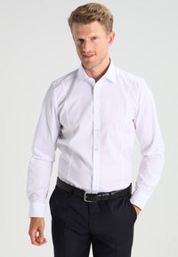 Weißes Langarmhemd mit Knopfleiste, spitzem Kragen und dezenten Nahtdetails, kombiniert mit dunklen Hosen und einem schwarzen Gürtel.