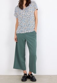Blusa de manga corta con estampado floral en azul, crema y verde, combinada con pantalones anchos verdes y sueltos, y mocasines negros con acentos dorados.