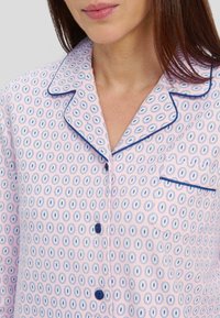 Camisa de pijama en rosa claro, con un patrón circular en azul y blanco, con cuello, bolsillo y botones azules. Textura de tela suave.