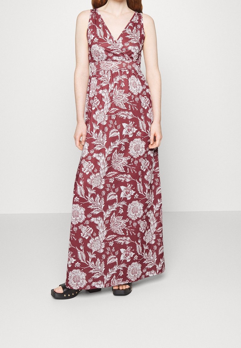 Femme portant une robe longue sans manches couleur bordeaux à motif floral blanc et des sandales à plateforme noires, debout devant un fond uni.