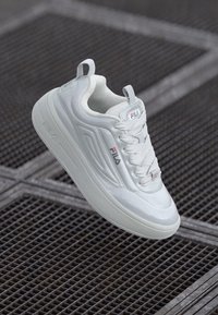 White sneaker with a smooth synthetic upper, rounded toe, and thick sole. Features Fila logo and textured accents on the side.