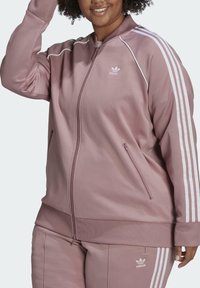 Chaqueta deportiva con cremallera color rosa claro con detalles blancos y tres rayas en las mangas. Cuenta con bolsillos laterales y dobladillo acanalado. Hecha de tela suave.