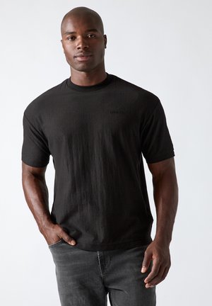 Mann trägt schwarzes T-Shirt mit kurzen Ärmeln und dunkle Jeans, steht vor einem einfarbigen hellen Hintergrund, eine Hand in der Tasche.