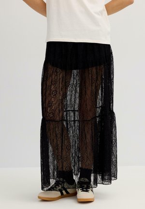 Falda maxi de encaje negro con diseño de volantes, textura transparente y una cintura elástica ancha, combinada con zapatillas de color claro.