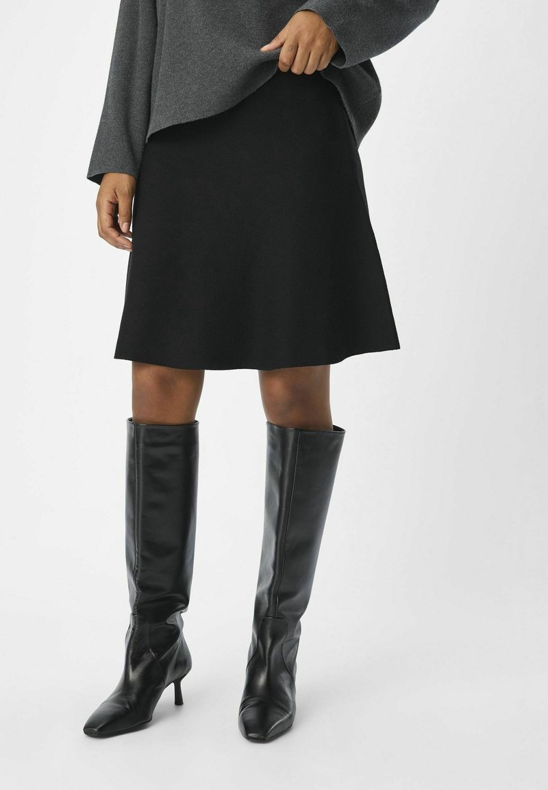 Femme portant une jupe noire trapèze, un pull gris et des bottes en cuir noires à talons arrivant aux genoux, debout devant un fond uni.