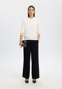 Weißer Sweatshirt mit Puffärmeln, kombiniert mit schwarzen, weit geschnittenen Hosen. Accessoires sind eine schwarze Clutch mit einer Schleife. Schwarze, spitz zulaufende High Heels.