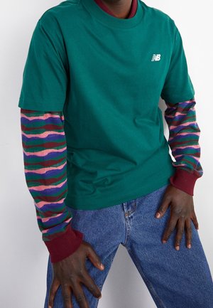 Grünes Baumwoll-T-Shirt, darüber ein Langarmshirt mit mehrfarbigen Wellenmustern. Denim-Jeans und bordeauxrote Bündchen komplettieren den Look.