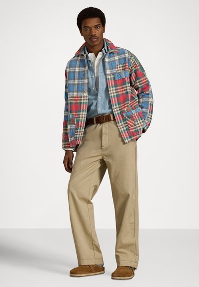 Καρό σακάκι σε μπλε, κόκκινο και πράσινο, φορεμένο πάνω από ένα τζιν πουκάμισο· το σύνολο ολοκληρώνουν μπεζ παντελόνια και καφέ παπούτσια χωρίς κορδόνια.