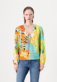 Blouse colorée et à motifs avec un col en V, des détails à cordon, des manches longues, et un mélange de teintes orange, verte, jaune et bleue.