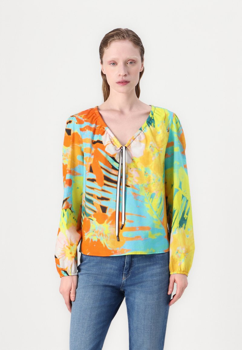 Blouse colorée et à motifs avec un col en V, des détails à cordon, des manches longues, et un mélange de teintes orange, verte, jaune et bleue.