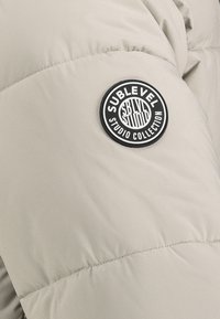 Beige gestepptes Material mit einem schwarz-weißen runden Aufnäher, auf dem "Sublevel Studio Collection" steht.