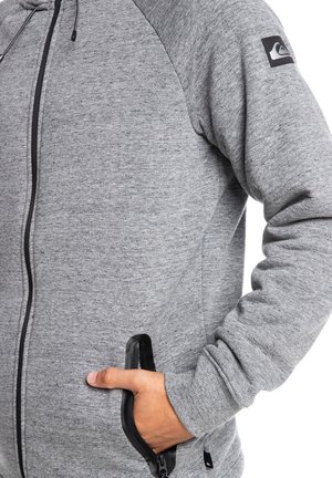 Grijze zip-up hoodie met een zachte textuur, zwarte accenten en een logopatch op de mouw. Zijzakken met ritsen voor veilige opslag.