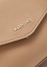 Bežová kožená kabelka s texturovaným povrchem, zdobená zlatým logem 'VALENTINO' a klopovým zapínáním se šitými detaily.