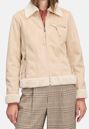 Veste en fausse peau de mouton beige avec poches à fermeture éclair et col, portée sur un haut couleur rouille et un pantalon taille haute à carreaux.
