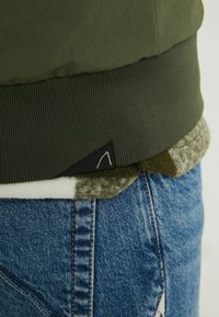 Olivově zelená bomber bunda s žebrovaným lemem, nasazená přes šedý a bílý vzorovaný svetr a modré džíny. Obsahuje malé černé logo.