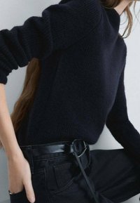 Dunkelblauer Strickpullover mit hohem Kragen und gerippten Bündchen, kombiniert mit schwarzen, hoch taillierten Hosen und einem schmalen schwarzen Gürtel.