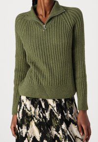 Suéter de punto acanalado verde oliva con cuello alzado y cierre de media cremallera. La tela tiene una textura suave y un corte relajado.