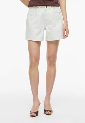 Personne portant un short blanc taille haute, un haut bordeaux et des mules marron ouvertes à talons, debout sur un fond blanc uni.