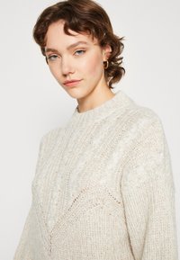 Lys beige strikket sweater med høj hals, der har et tekstureret mønster og subtile kabeldetaljer over fronten. Blød, afslappet stil.