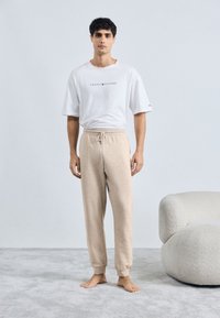 Pantaloni de trening bej deschis, cu margini cu manșete și talie cu șnur, asortați cu un tricou alb cu mânecă scurtă, având un imprimeu cu logo. Textură moale a materialului.