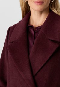 Donna che indossa un cappotto di lana viola scuro sopra una camicia coordinata con bottoni, mostrando il colletto, le rever e parzialmente il viso con capelli biondi.