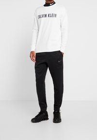 Muž v bílém tričku s dlouhým rukávem Calvin Klein, černém roláku Nike, černých teplácích Nike a černých teniskách Nike, stojící na bílém pozadí.