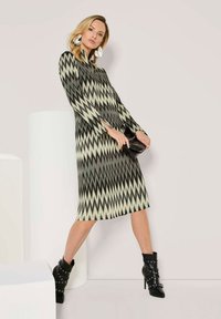 Frau in einem schwarzen, beigen, zickzackmuster-gehäkelten Midikleid, großen weißen Ohrringen, die eine schwarze Clutch hält, sowie schwarzen Stiefeletten mit Schnallen.