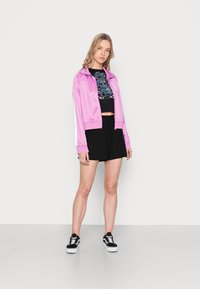 Light pink satin jacket with white side stripes, black cropped top featuring a blue graphic, and black shorts. Black and white sneakers.