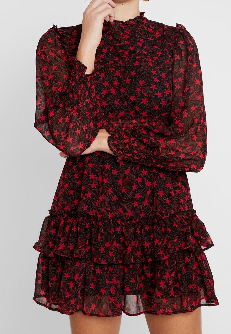 Femme portant une robe noire transparente avec des motifs d'étoiles rouges, manches longues bouffantes, poignets froncés, et jupe à volants en étages, main près du visage.