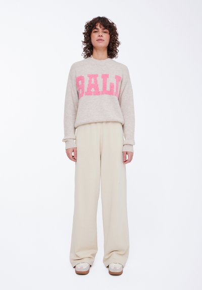 Beige strikket sweater med fed pink "BALI" tekst, afslappet pasform og ribbede manchetter, parret med brede cremefarvede bukser og lyse sneakers.