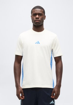 Giovane uomo nero di fronte che indossa una maglietta bianca con strisce laterali blu e un piccolo logo sportivo blu, abbinata a pantaloncini scuri.