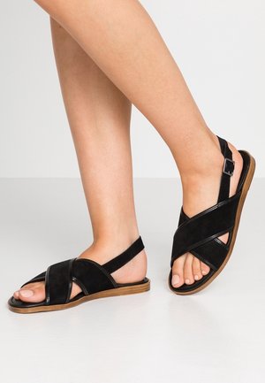 Des pieds portant des sandales en suède noir à brides croisées avec des semelles marron, un pied légèrement levé montrant une bride ajustable.
